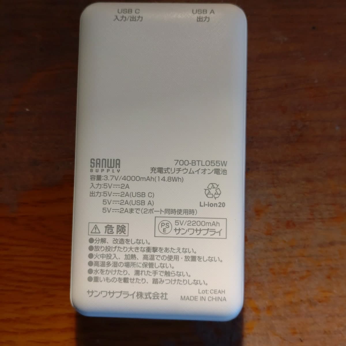 木の机に置かれた、白くて角の丸いモバイルバッテリーの裏面。上端にUSB C 入力/出力とUSB A 出力の文字、中ほどにSANWA SUPPLY、700-BTL055W、容量3.7V/4000mAhと印刷されている