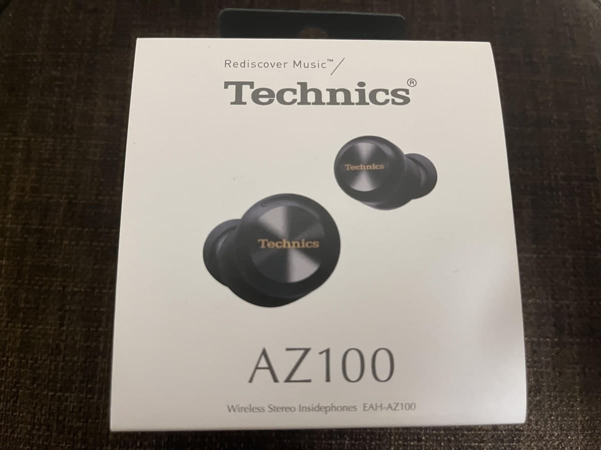 濃い茶色の布の上に置かれた、封を切っていない白い箱。Technicsのロゴと黒いイヤホンの写真、AZ100という大きな文字、Wireless Stereo Insidephones EAH-AZ100の表記が並んでいる