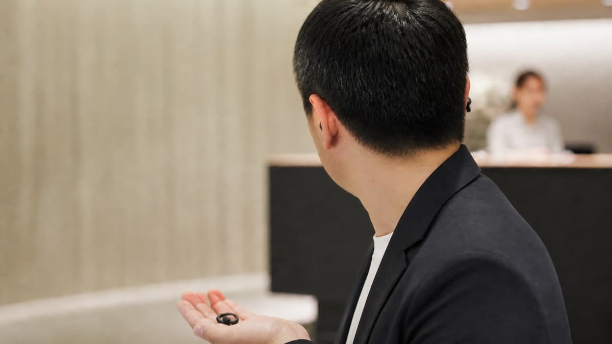 受付カウンターの前で、片方のイヤホンを耳から外して手のひらに載せ、カウンターのほうへ顔を向けている人の胸から上