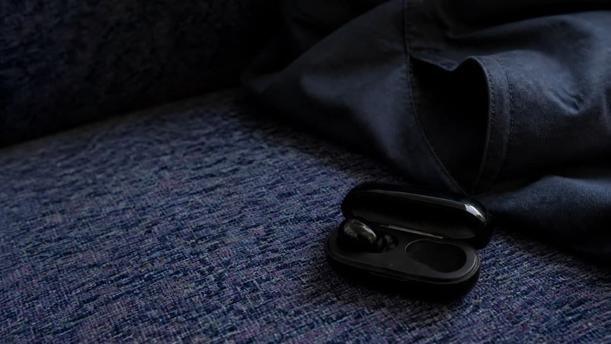 電車の布張りの座席の上に転がった、つやのある黒い充電ケース。ふたが少し開いていて、中の2つの窪みのうち片方にしかイヤホンが入っていない。すぐ上に上着の裾とポケットの口が写っている