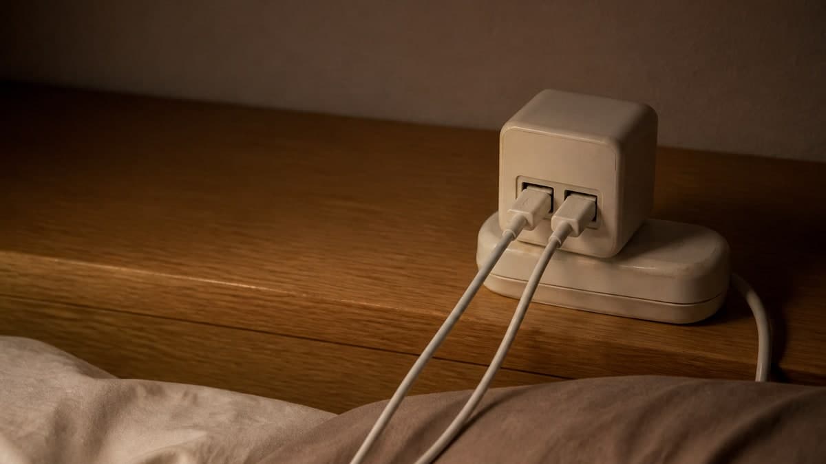夜の寝室のベッドボードの木の面に置かれた、差込口が2つある白いUSB充電器。画面の下のふちには毛布のふちが写り、充電器はそこから少し離して置かれている。ケーブルが2本手前へ伸びている