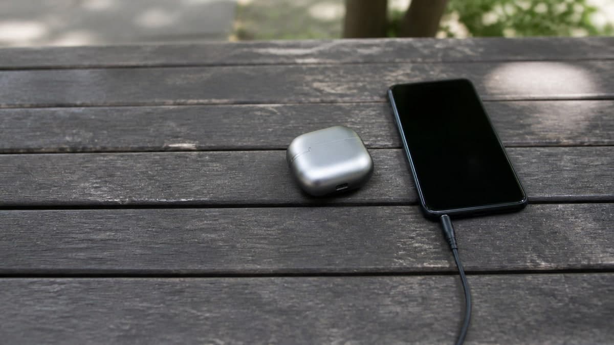屋外のベンチの木の座面に置かれた、ふたを閉じた銀色のワイヤレスイヤホンの充電ケース。その隣に画面を消したスマートフォンがあり、有線イヤホンのケーブルが挿さって手前へ伸びている
