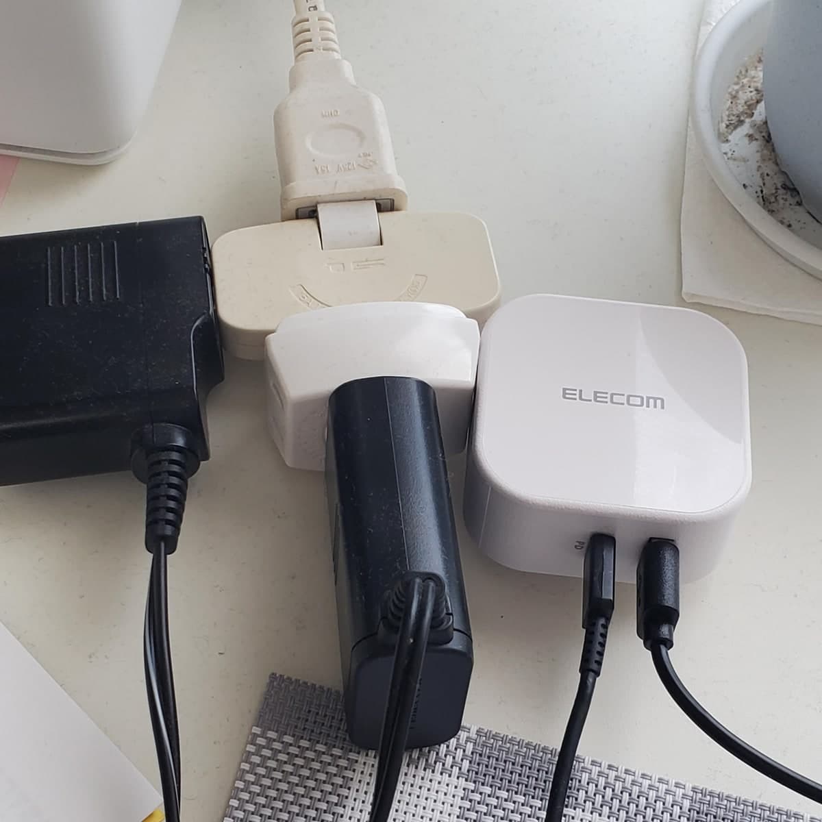 白い机の上の電源タップに、ELECOMと書かれた白い四角い充電器が挿してある。黒いアダプタ2つと並んでいて、充電器からは黒いケーブルが2本のびている