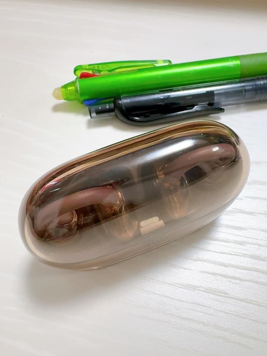 白い机に置かれた、中身が透けて見える小判形の充電ケース。うすいピンク色のイヤホンが2つ、輪の形に丸まって入っている。奥に緑と黒のボールペンが2本置いてあり、ケースがペンの半分ほどの長さだと分かる