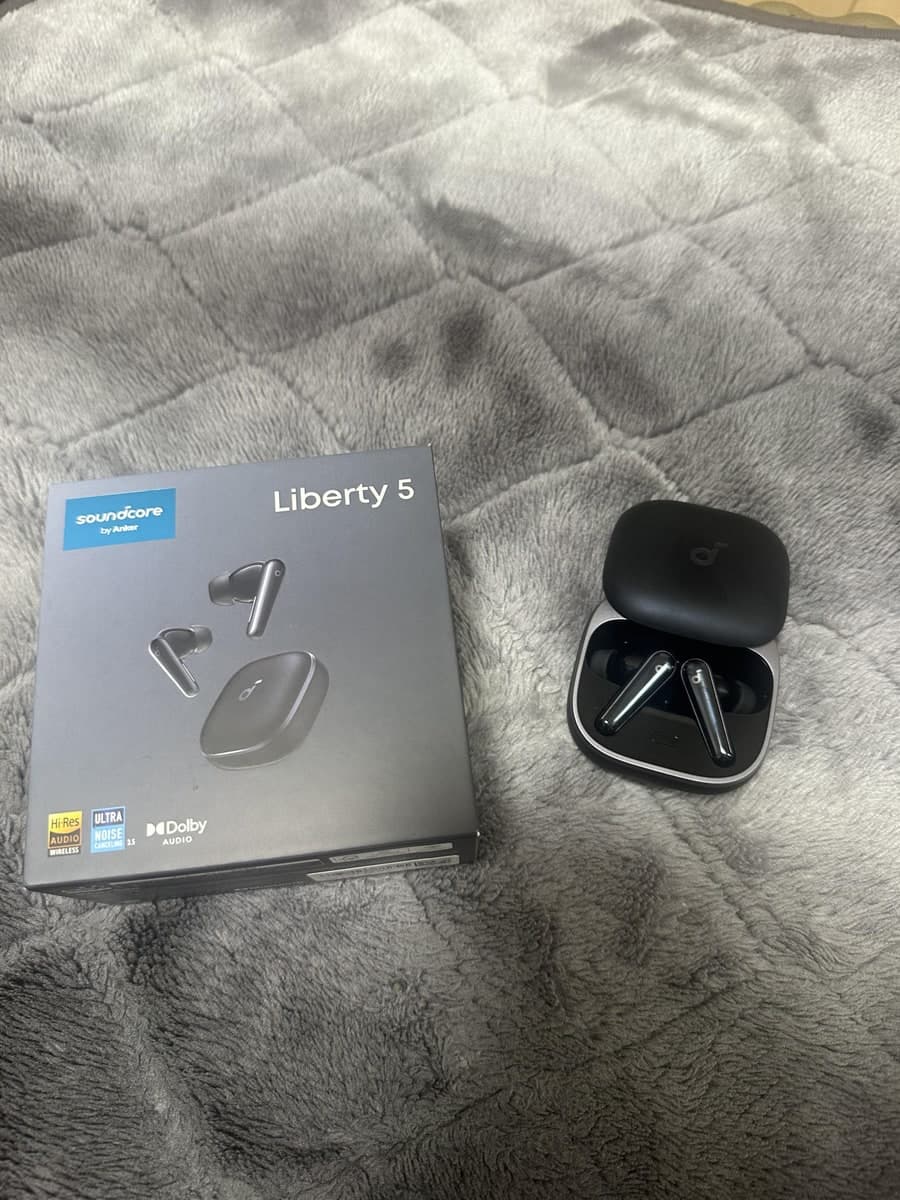 灰色の毛布の上に、soundcore Liberty 5と書かれた黒い箱と、フタを開けた黒い充電ケースが並べて置かれている。ケースの中には黒いイヤホンが2つ入っている