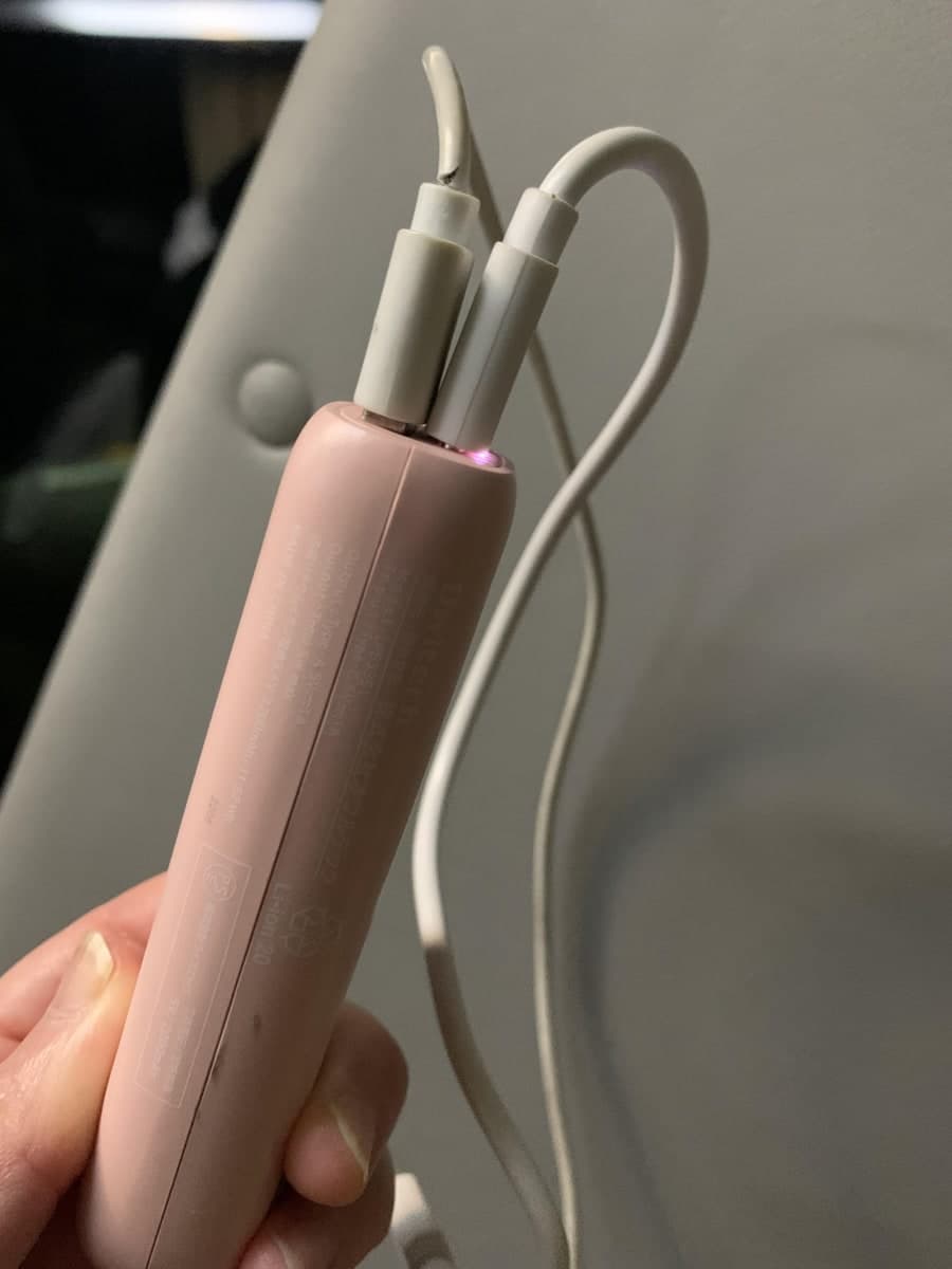 手に持った、うすいピンク色の細長い棒のようなモバイルバッテリー。上の端に白いケーブルが2本挿さっていて、その根元のランプが紫色に光っている