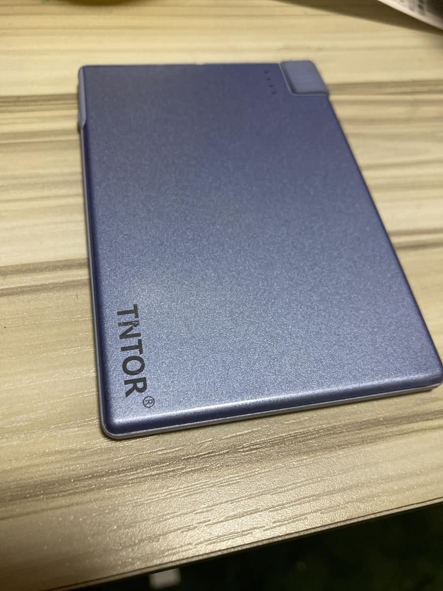 木目の机に置かれた、カードのように薄くて平たい紫色のモバイルバッテリー。表面にTNTORのロゴがあり、上の端に残量を示す4つの小さなランプと、短い突起が並んでいる